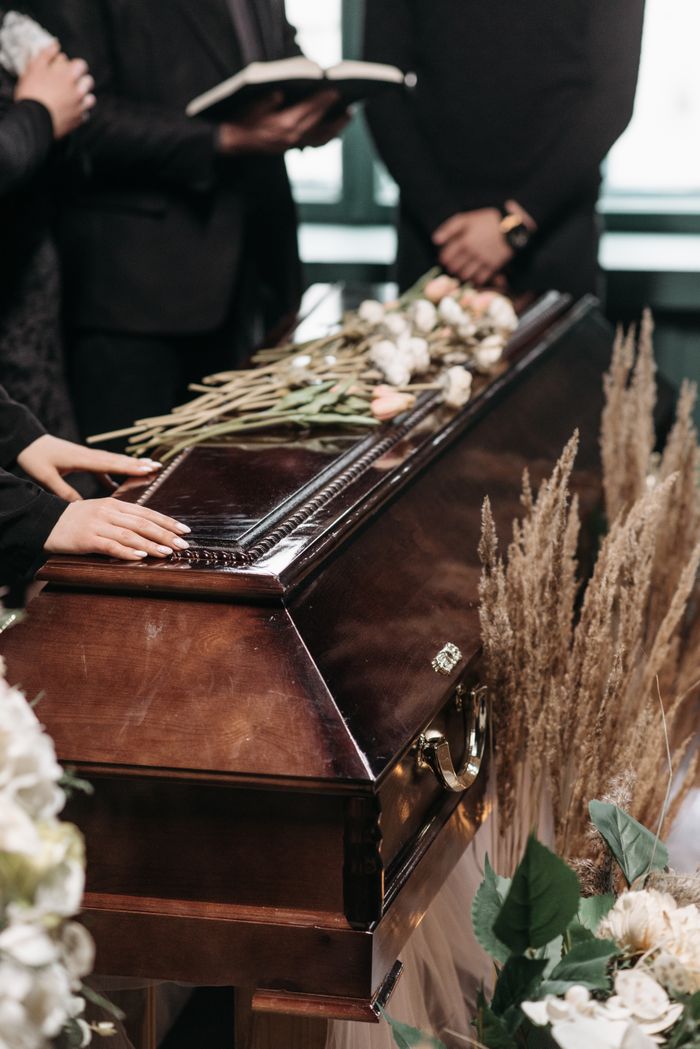 creative-approach-img Mourners at a funeral with floral arrangements on a polished wooden coffin.